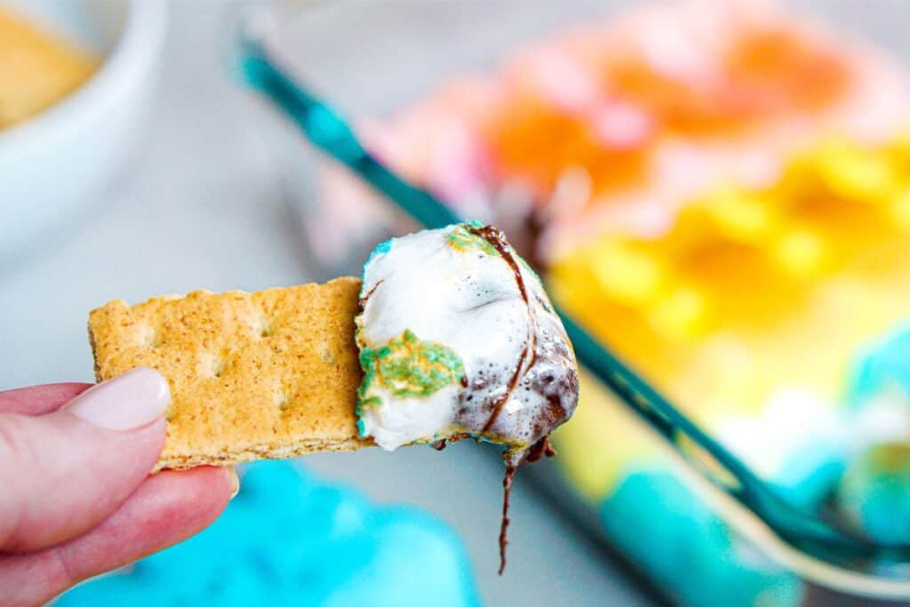 Close-up of a hand holding a graham cracker dipped in melted, multicolored marshmallow with chocolate, reminiscent of a festive S'mores Dip. A tray of vibrant Easter Bunny-inspired Peeps marshmallows is blurred in the background.