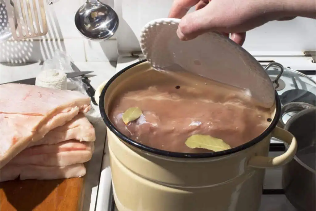 Person placing a lid on a pot of simmering chicken with bay leaves. Slices of pork fat, perfect for traditional meat storage without a refrigerator, lie on a cutting board nearby. Kitchen utensils in the background complete the cozy culinary scene.