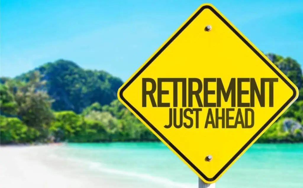 Yellow road sign with "Retirement Just Ahead" text stands against a tropical beach backdrop, where clear blue waters and lush green foliage invite you to plan your perfect escape now.