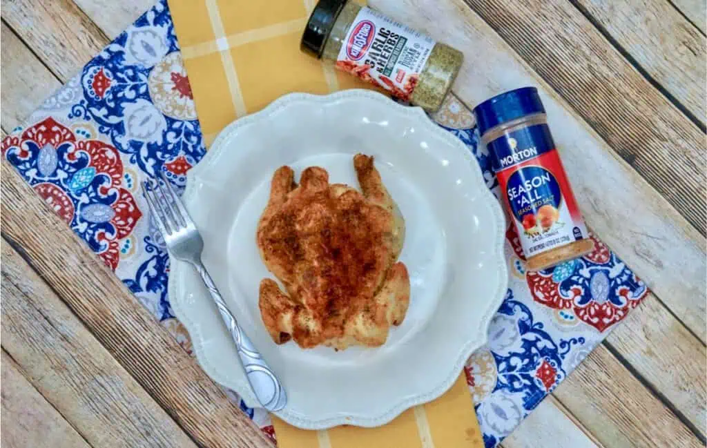A beautifully roasted Cornish hen rests on a plate with a fork, sitting atop a patterned napkin. Flanked by two spice containers on the wooden table, it showcases how air fryer magic can create an exquisite dining experience.