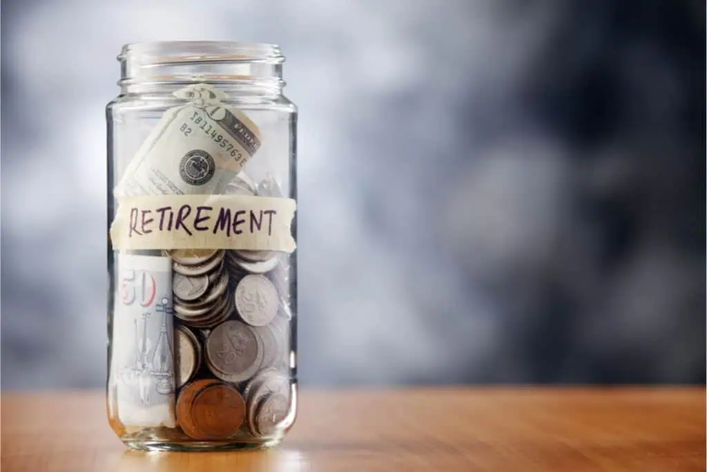 A jar labeled "retirement" overflows with a mix of coins and paper money, sitting on a wooden surface, symbolizing the fruits of careful retirement planning.
