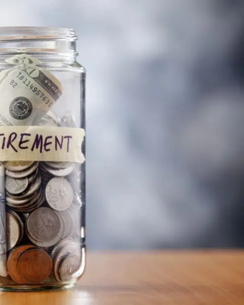 A jar labeled "retirement" overflows with a mix of coins and paper money, sitting on a wooden surface, symbolizing the fruits of careful retirement planning.
