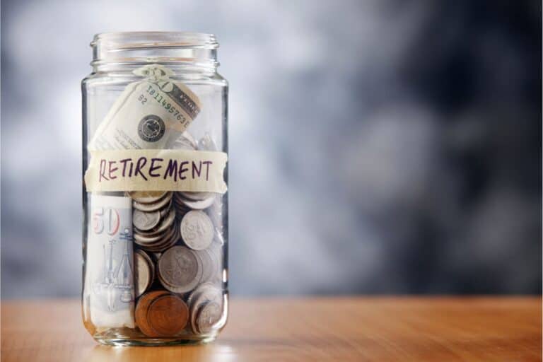 A jar labeled "retirement" overflows with a mix of coins and paper money, sitting on a wooden surface, symbolizing the fruits of careful retirement planning.