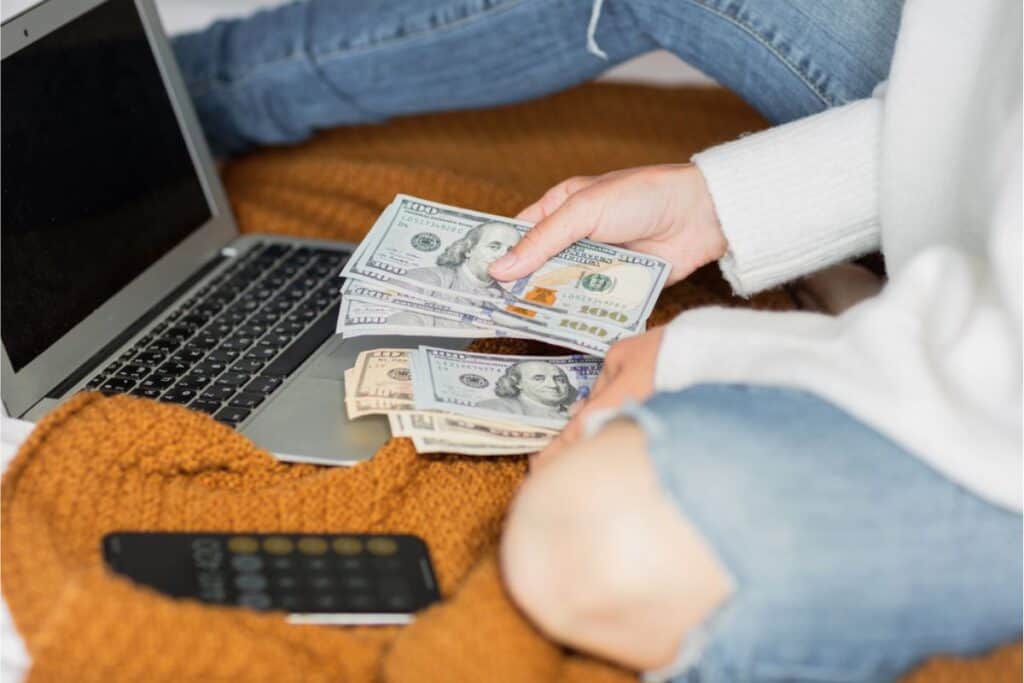 A person sits on a bed with a laptop open to an internet page about how to save money, counting various denominations of U.S. dollar bills. A smartphone lies nearby on a brown blanket, possibly ready for the next tip or financial app download.