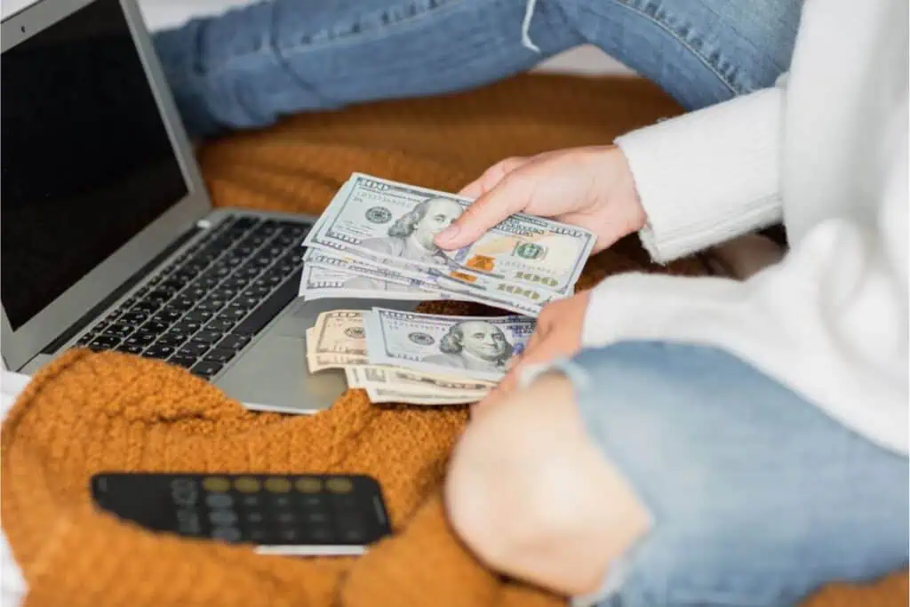 A person sits on a bed with a laptop open to an internet page about how to save money, counting various denominations of U.S. dollar bills. A smartphone lies nearby on a brown blanket, possibly ready for the next tip or financial app download.
