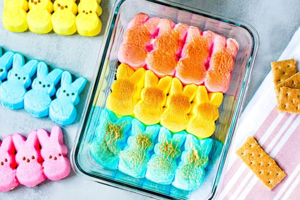 Colorful marshmallow bunnies, reminiscent of Peeps, are artfully arranged in a glass container, with extra pink and yellow bunnies nearby. Graham crackers sit invitingly on a striped cloth to the side, perfect for crafting an Easter Bunny-themed s’mores dip.