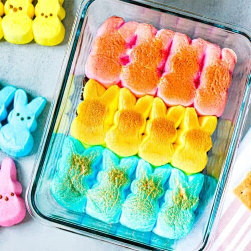 Colorful marshmallow bunnies, reminiscent of Peeps, are artfully arranged in a glass container, with extra pink and yellow bunnies nearby. Graham crackers sit invitingly on a striped cloth to the side, perfect for crafting an Easter Bunny-themed s’mores dip.