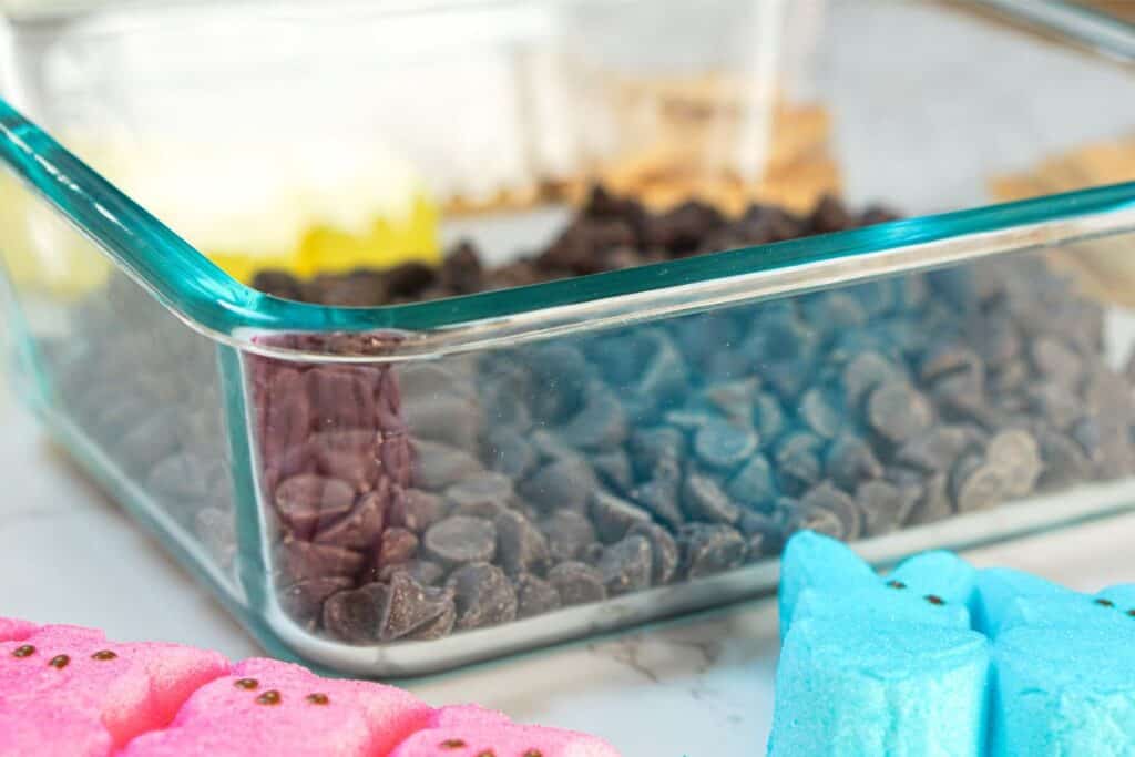 A glass dish filled with chocolate chips is surrounded by vibrant marshmallow Peeps, creating a delightful scene reminiscent of Easter Bunny festivities on a marble surface.