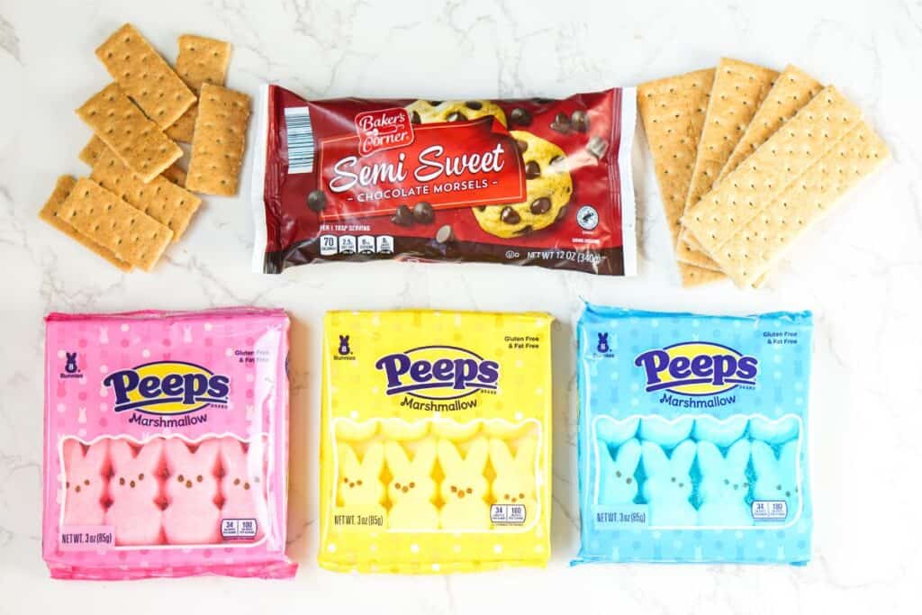 Packages of pink, yellow, and blue Peeps marshmallows alongside a bag of semisweet chocolate chips and graham crackers on a marble surface—perfect ingredients for an Easter Bunny-inspired S'mores Dip.