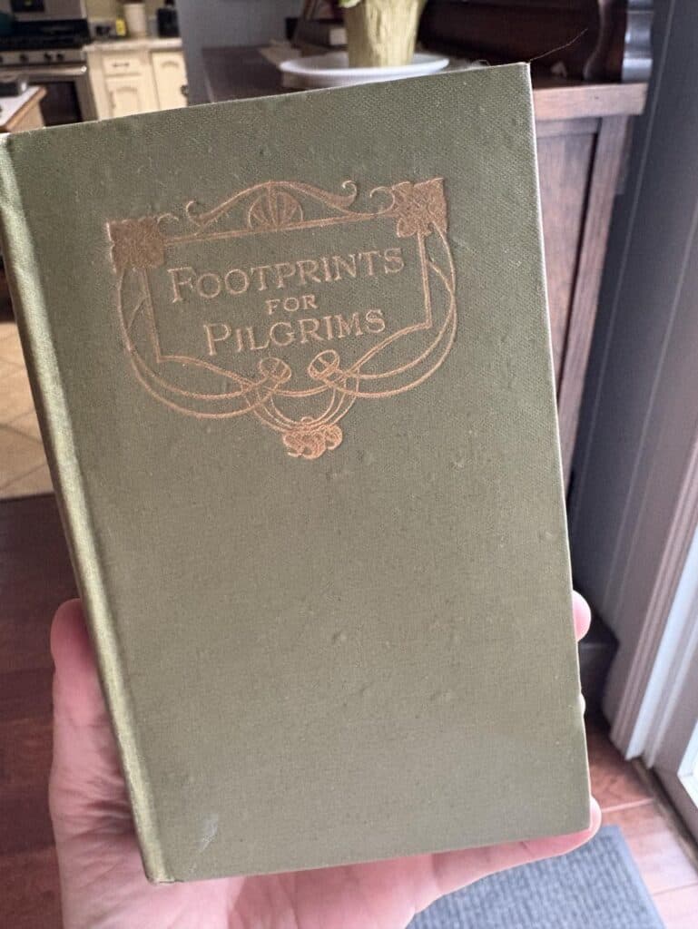 A person holds a green hardcover book titled "Footprints for Pilgrims," its cover shining with ornate gold lettering as if kissed by sunshine.