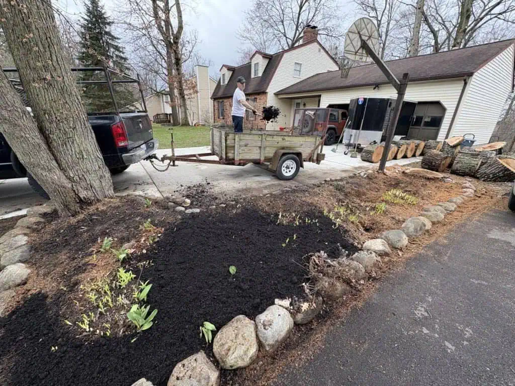 A person personally posts their progress as they unload landscaping materials from a trailer in the driveway. Nearby, a basketball hoop stands tall, with chopped logs scattered around. Young plants flourish in a mulch bed bordered by rocks, framed by trees and the house beyond.