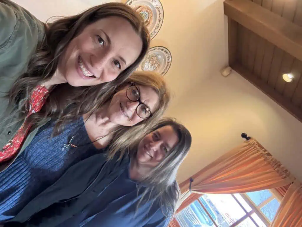 Three women smiling, standing indoors near a window with orange curtains that let in streams of sunshine. Ceiling beams and decorative plates add charm to the background.