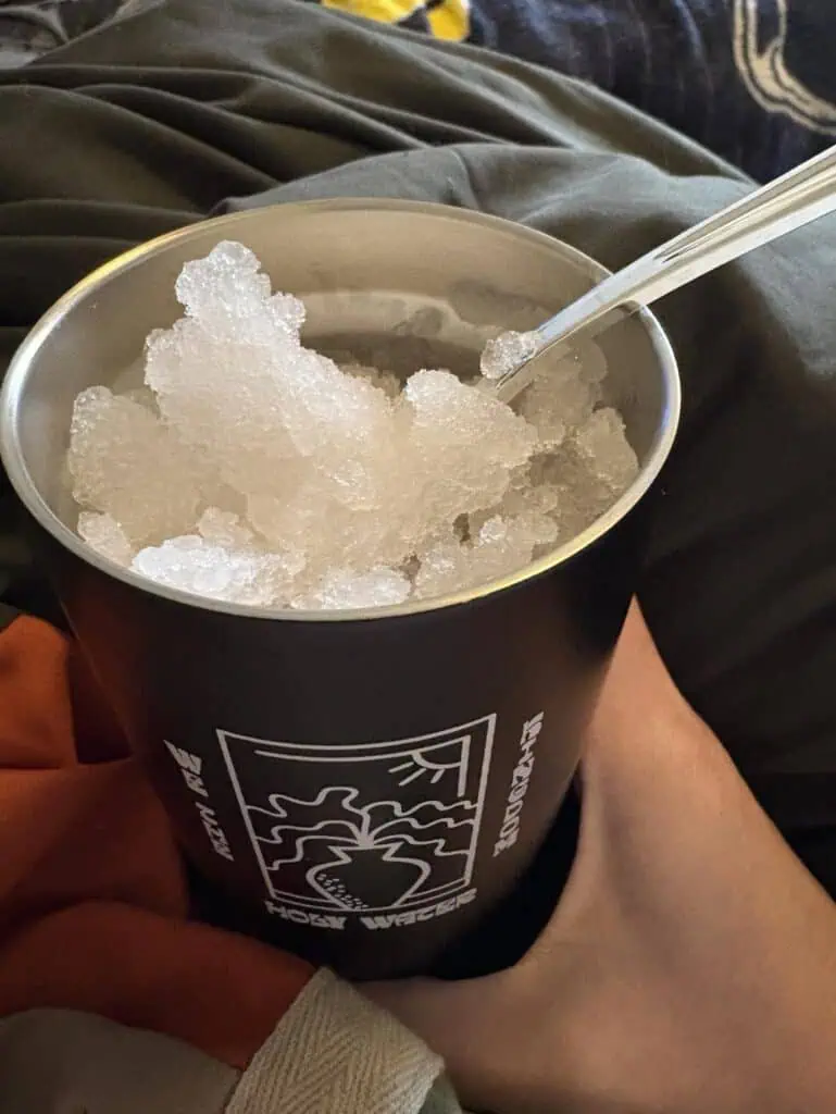 A personal touch is captured as a hand holds an aluminum cup filled with crushed ice and a spoon, set against a dark fabric background.
