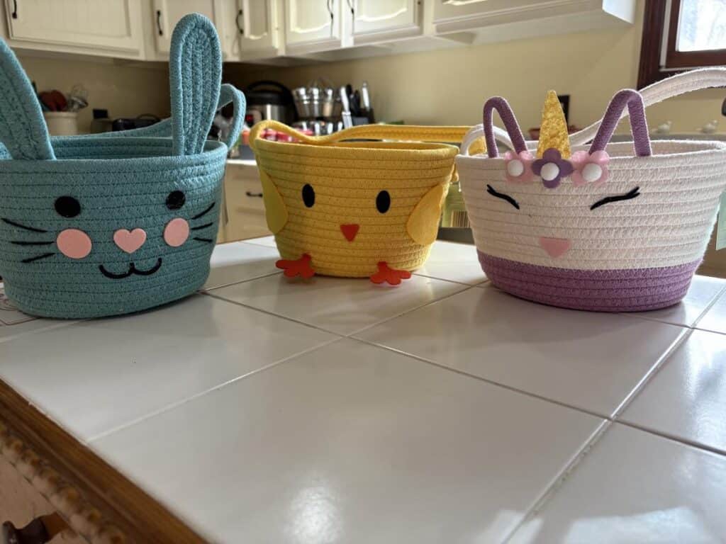 Three woven baskets on a countertop: a blue bunny, a yellow chick, and a white unicorn with pink accents, bringing sunshine to the kitchen in the background.