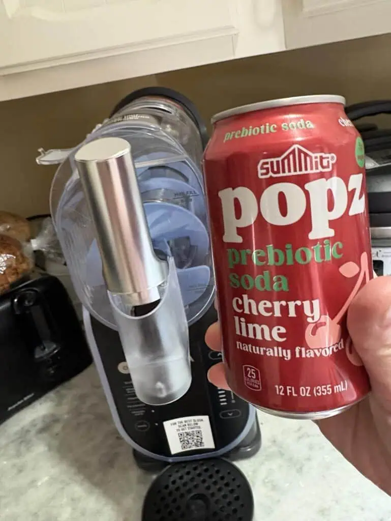 A hand casually holds a can of Summit Popz prebiotic soda, cherry lime flavor, next to a trusty soda maker on the kitchen counter. Perfect for sharing in a personal post about vibrant flavors and home-brewed fizz delight.
