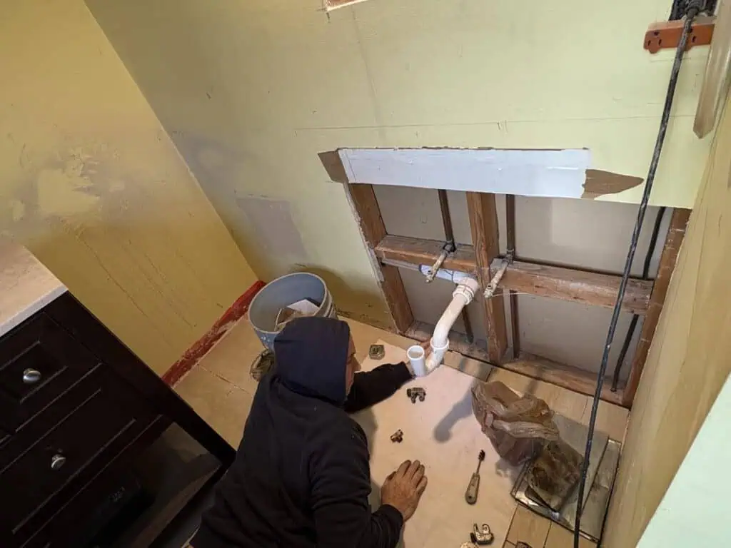 In a partially remodeled room, a person in a hoodie is immersed in plumbing tasks under the sink, surrounded by an array of tools and parts.
