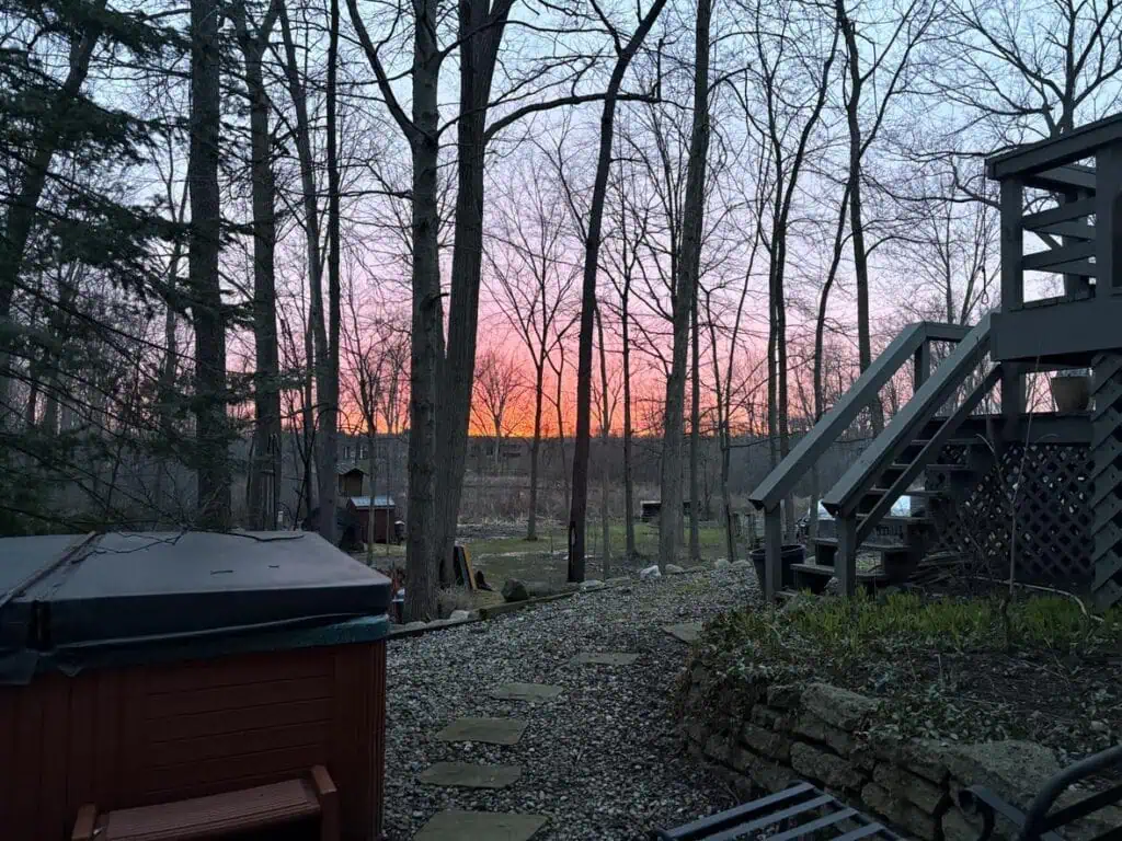 Backyard with trees and a gravel path at sunset, perfect for personal blog inspiration. A hot tub and deck stairs are on the right beneath a stunning pink and orange sky—blog content just waiting to be shared.