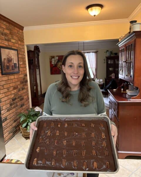 A woman beams with joy as she holds a large tray of chocolate dessert with pecan toppings in a beautifully themed kitchen setting, highlighting her personal touch.