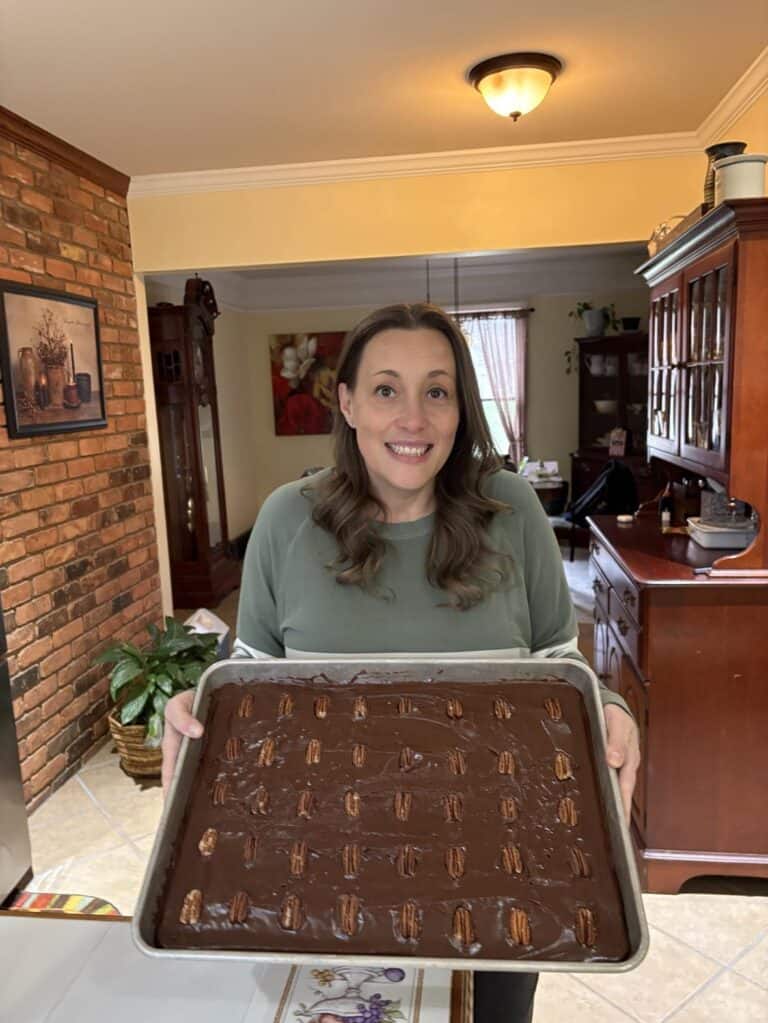 A woman beams with joy as she holds a large tray of chocolate dessert with pecan toppings in a beautifully themed kitchen setting, highlighting her personal touch.
