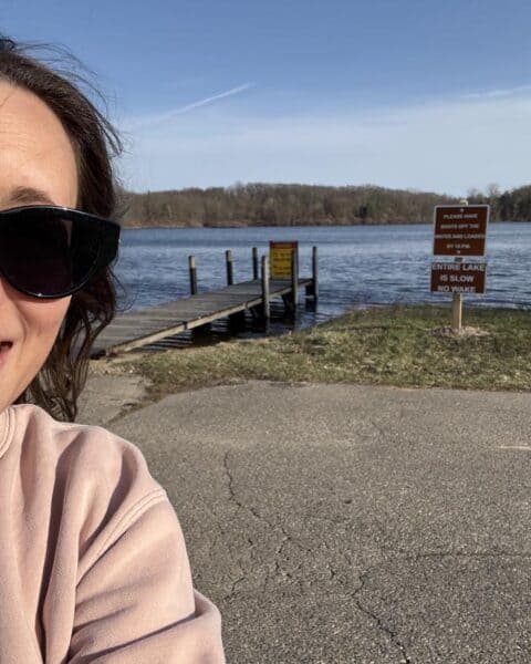 Personal post: Woman wearing sunglasses smiles in front of a lake with a wooden dock; informational signs are posted near the water’s edge. Trees and blue sky create a serene background.