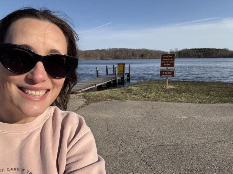 Personal post: Woman wearing sunglasses smiles in front of a lake with a wooden dock; informational signs are posted near the water’s edge. Trees and blue sky create a serene background.