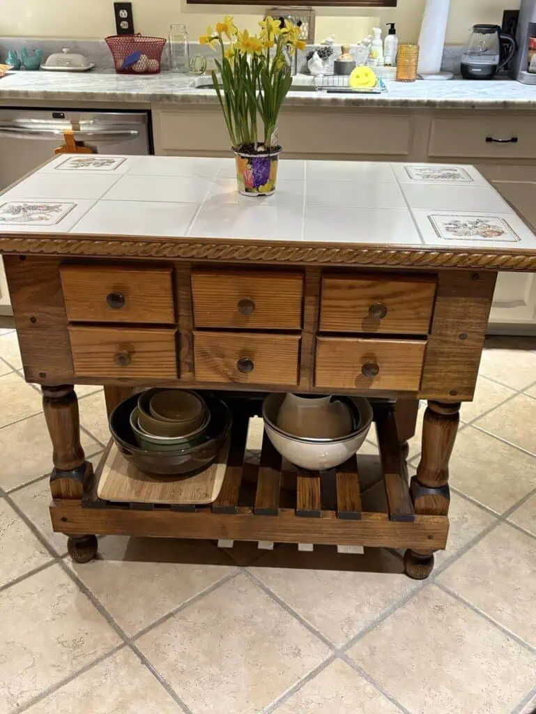 The kitchen island, styled with personal charm, showcases a wooden structure complete with drawers and shelf. It boasts a tiled countertop adorned by a pot of yellow flowers, while bowls and plates find their designated post on the lower shelf.