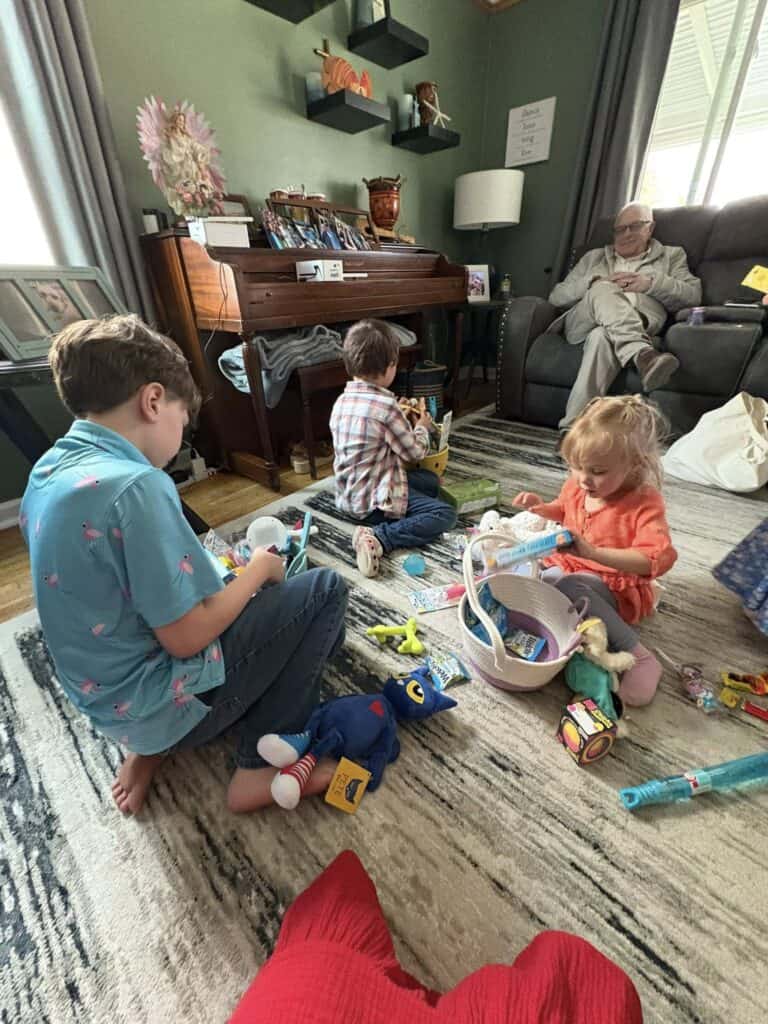 Three children sit on a living room rug sorting through a basket of toys and candy, while an elderly man relaxes in an armchair in the background.