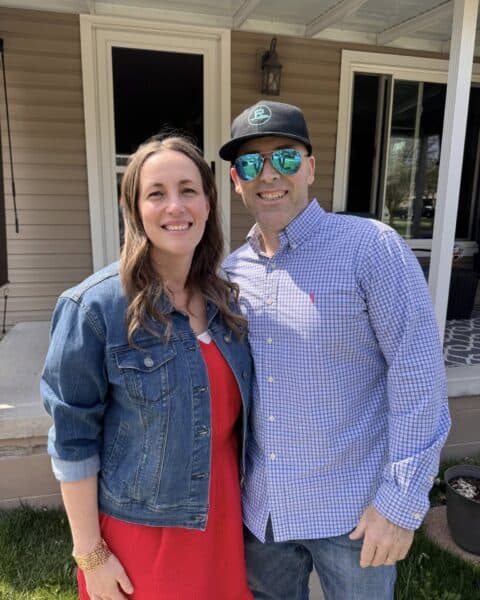 A woman in a red dress and denim jacket stands next to a man in a checked shirt, sunglasses, and black cap, both smiling outside a house—capturing a joyful moment in this personal post.