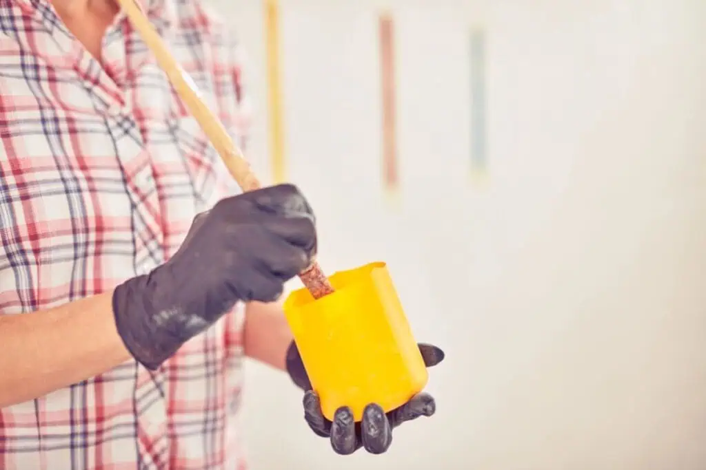 Person wearing black gloves stirs paint in a yellow container with a wooden stick, wearing a plaid shirt. Vertical color swatches adorn the white wall, hinting at creative ways to save money on home improvements.
