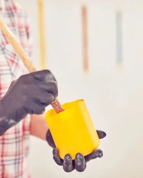 Person wearing black gloves stirs paint in a yellow container with a wooden stick, wearing a plaid shirt. Vertical color swatches adorn the white wall, hinting at creative ways to save money on home improvements.
