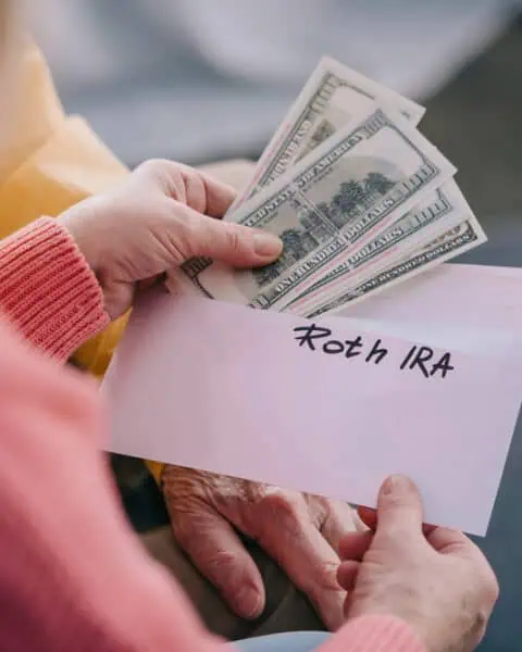 A person holds cash and an envelope labeled "Roth IRA," suggesting depositing money into an individual retirement arrangement or IRA for future savings.