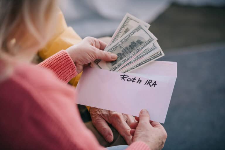 A person holds cash and an envelope labeled "Roth IRA," suggesting depositing money into an individual retirement arrangement or IRA for future savings.