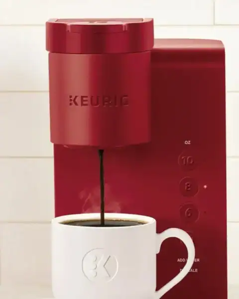 A vibrant red Keurig coffee maker dispenses rich coffee into a white mug, accompanied by a spoon. A coffee pod holder filled with assorted pods sits to the left against a pristine white tile backsplash, offering endless choices for your perfect brew.