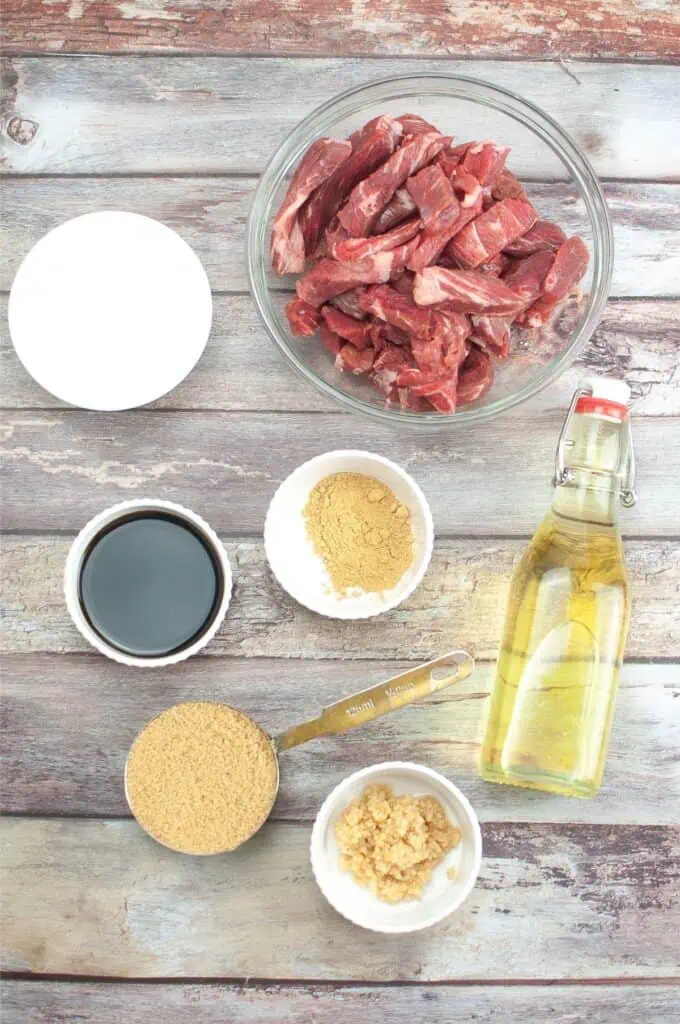 Ingredients for a slow cooker Mongolian Beef recipe: sliced beef in a bowl, soy sauce, brown sugar, ground ginger, and minced garlic in bowls, all beside a bottle of oil on a wooden surface.