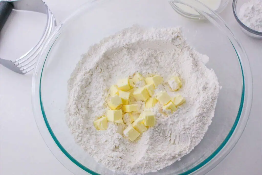 A glass bowl with flour and cubed butter rests on a white surface, ready for a scone recipe. It's accompanied by a pastry cutter and a small bowl of milk, reminiscent of classic Bisquick magic.