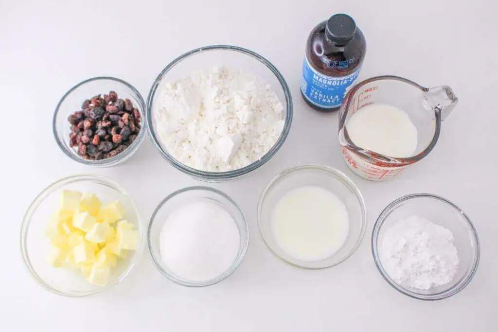 Seven bowls with baking ingredients: raisins, flour, butter, sugar, milk, vanilla extract, and powdered sugar rest on a white surface—everything you need for an easy recipe to whip up homemade scones.