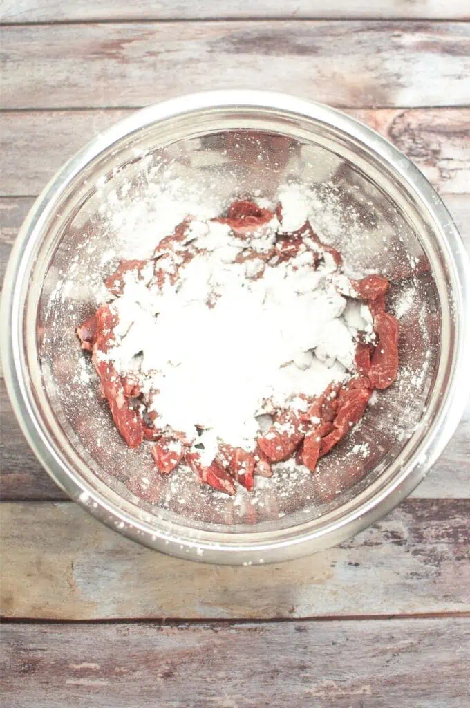 A glass bowl containing strips of raw meat coated with white flour rests on a wooden surface, ready to be transformed into a savory Mongolian beef recipe.