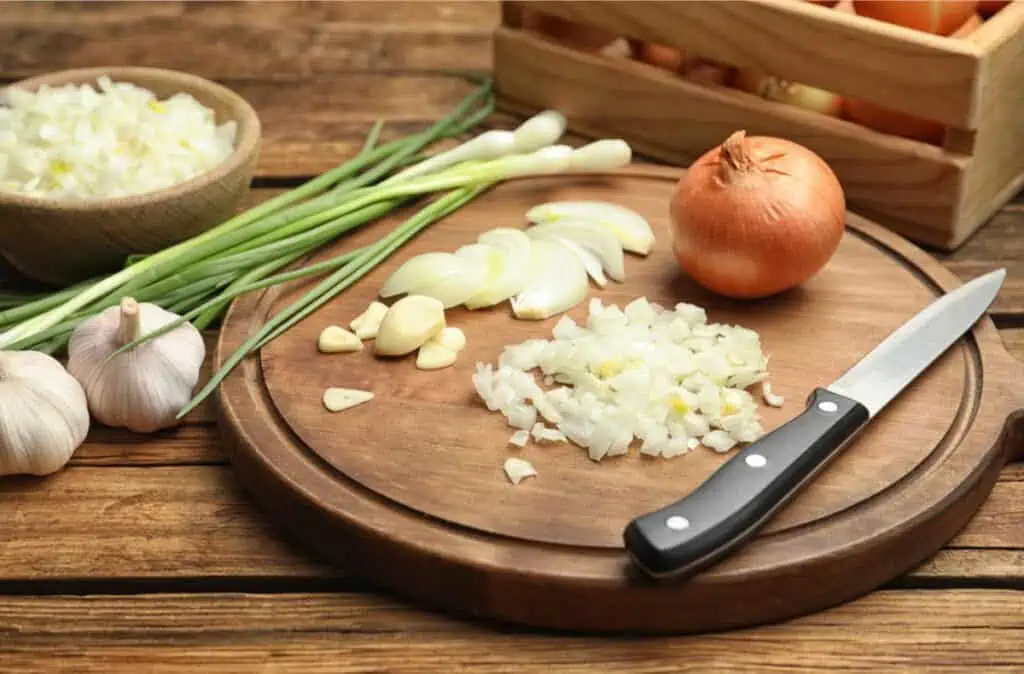 Chopped onions on a wooden cutting board with a knife, surrounded by whole onions, garlic, and green onions on a wooden table—with clever tricks to cut onions without crying.
