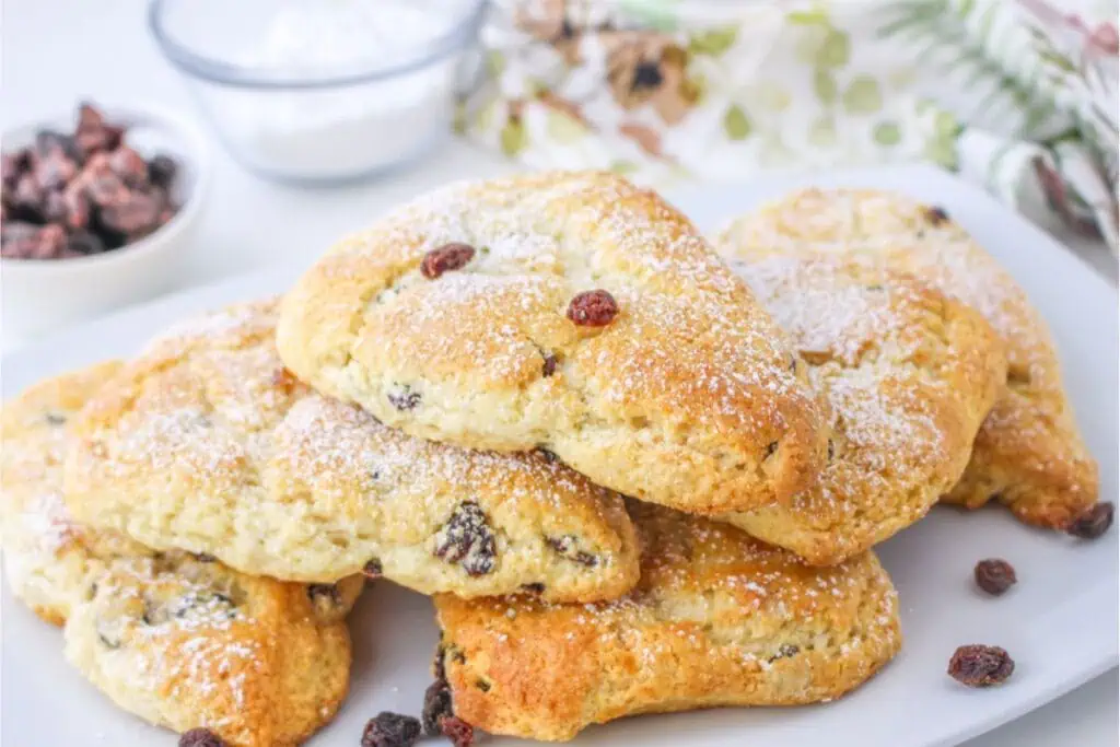 A plate of four triangular raisin scones, crafted from a classic Bisquick recipe and dusted with powdered sugar. A small bowl of raisins and a floral napkin provide the perfect backdrop.