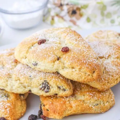 A plate of four triangular raisin scones, crafted from a classic Bisquick recipe and dusted with powdered sugar. A small bowl of raisins and a floral napkin provide the perfect backdrop.