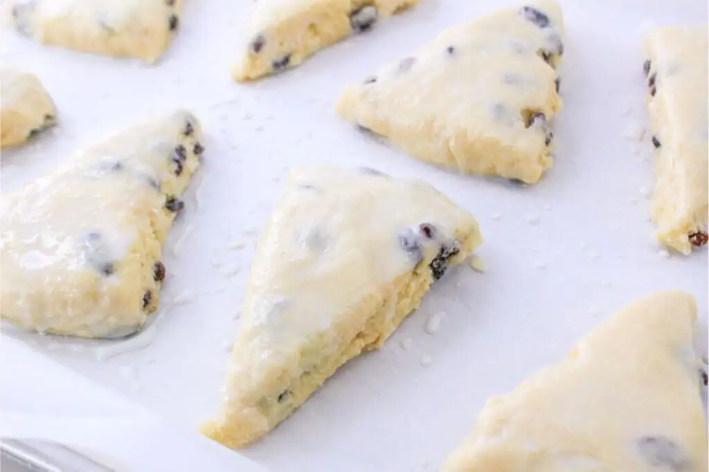Triangular scones with a light glaze and visible raisins are placed on a baking sheet lined with parchment paper, showcasing an easy and delicious twist on the classic scone using a Bisquick recipe.