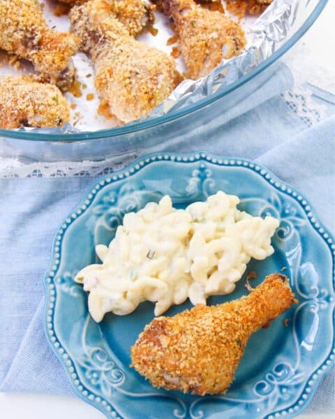 A blue plate with a breaded chicken drumstick and macaroni salad showcases the taste of homemade shake and bake. More drumsticks are tempting on a foil-lined tray in the background.