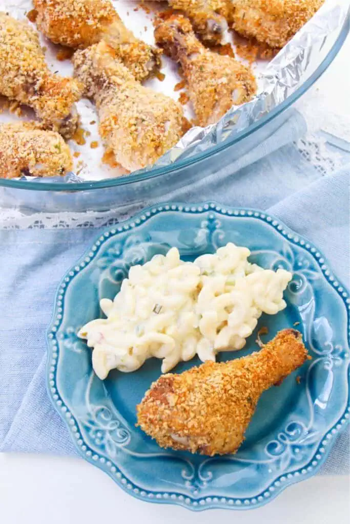 A blue plate with a breaded chicken drumstick and macaroni salad showcases the taste of homemade shake and bake. More drumsticks are tempting on a foil-lined tray in the background.