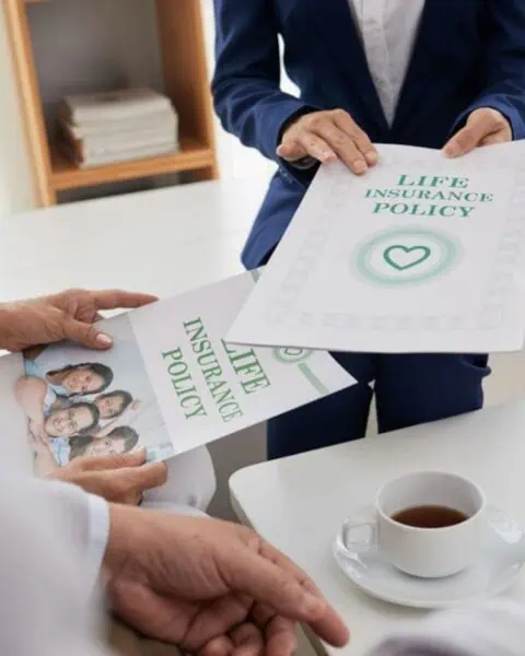 Two people discuss how to buy life insurance as they exchange policy documents at a desk with a laptop and a cup of coffee.