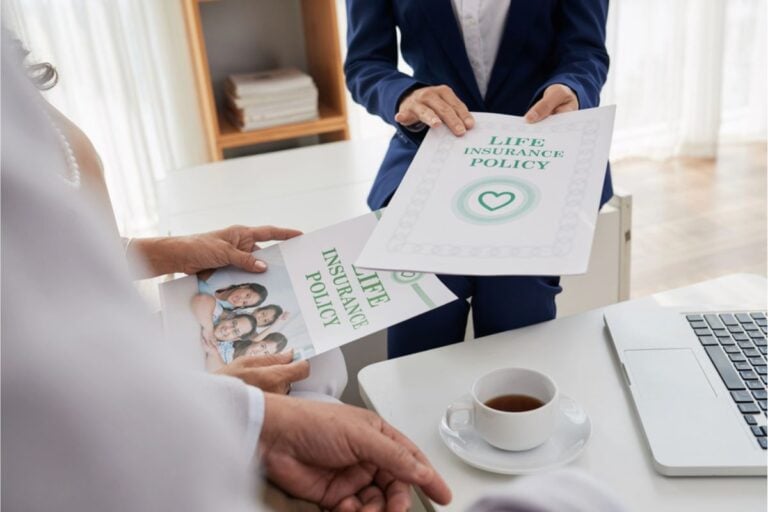 Two people discuss how to buy life insurance as they exchange policy documents at a desk with a laptop and a cup of coffee.