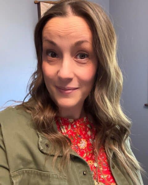A woman with long brown hair, wearing a floral red top and green jacket, stands indoors. Her smile radiates warmth like a touch of sunshine, capturing the heart effortlessly as she gazes at the camera.