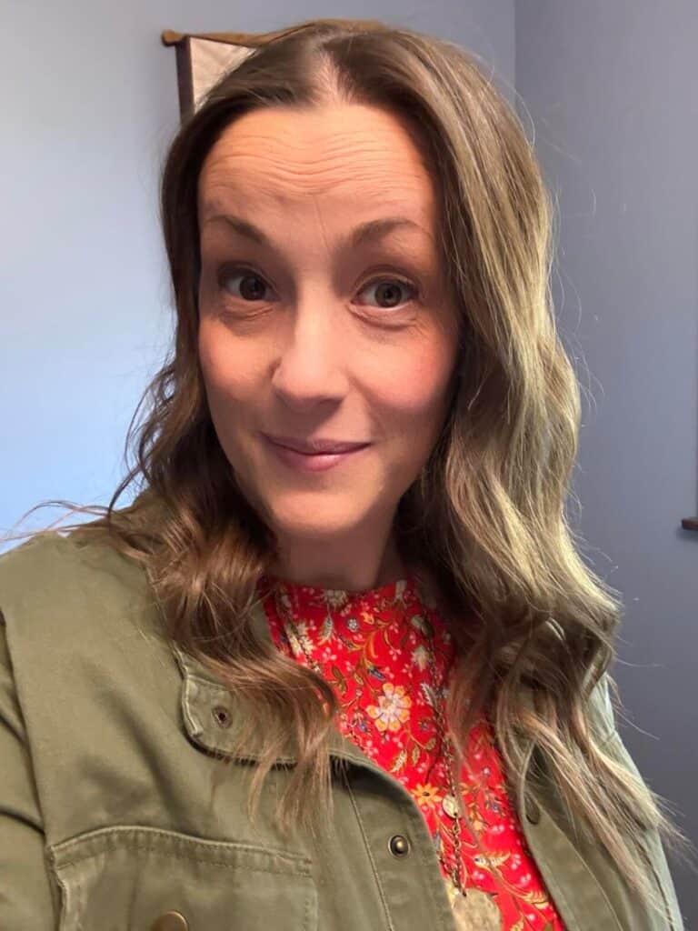 A woman with long brown hair, wearing a floral red top and green jacket, stands indoors. Her smile radiates warmth like a touch of sunshine, capturing the heart effortlessly as she gazes at the camera.