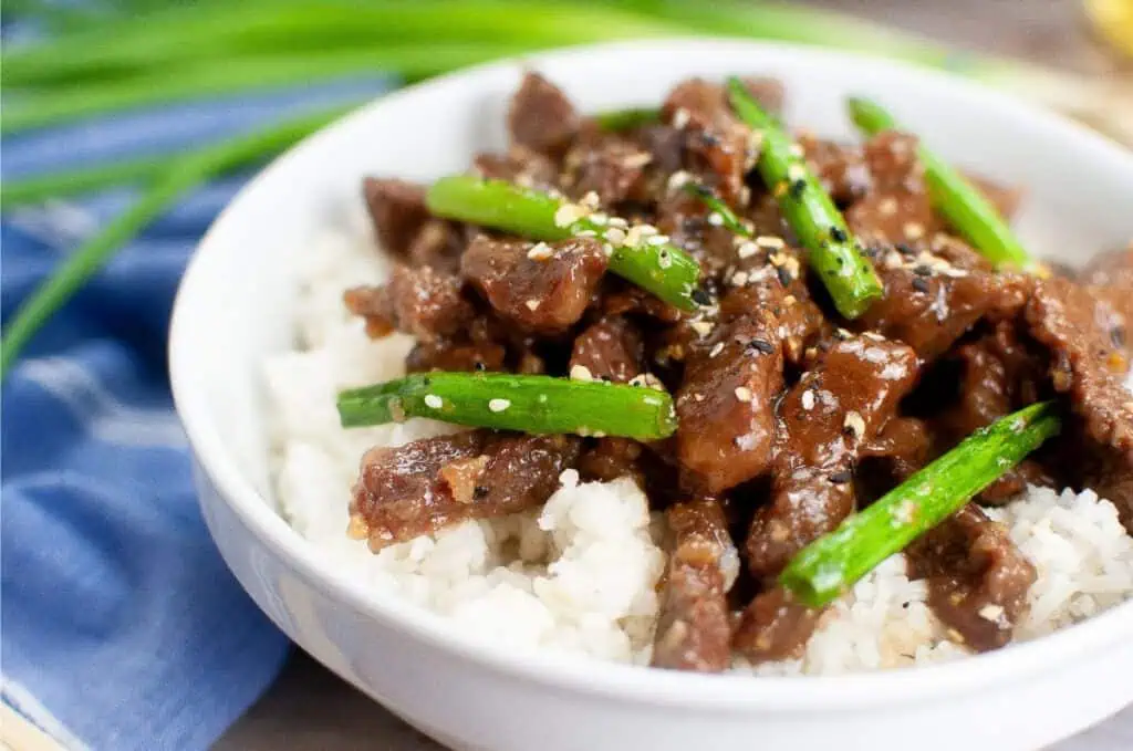 A bowl of savory Mongolian Beef features tender slices of beef and green onions over rice, garnished with sesame seeds.