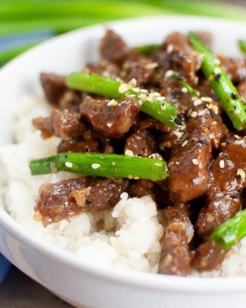 A bowl of savory Mongolian Beef features tender slices of beef and green onions over rice, garnished with sesame seeds.