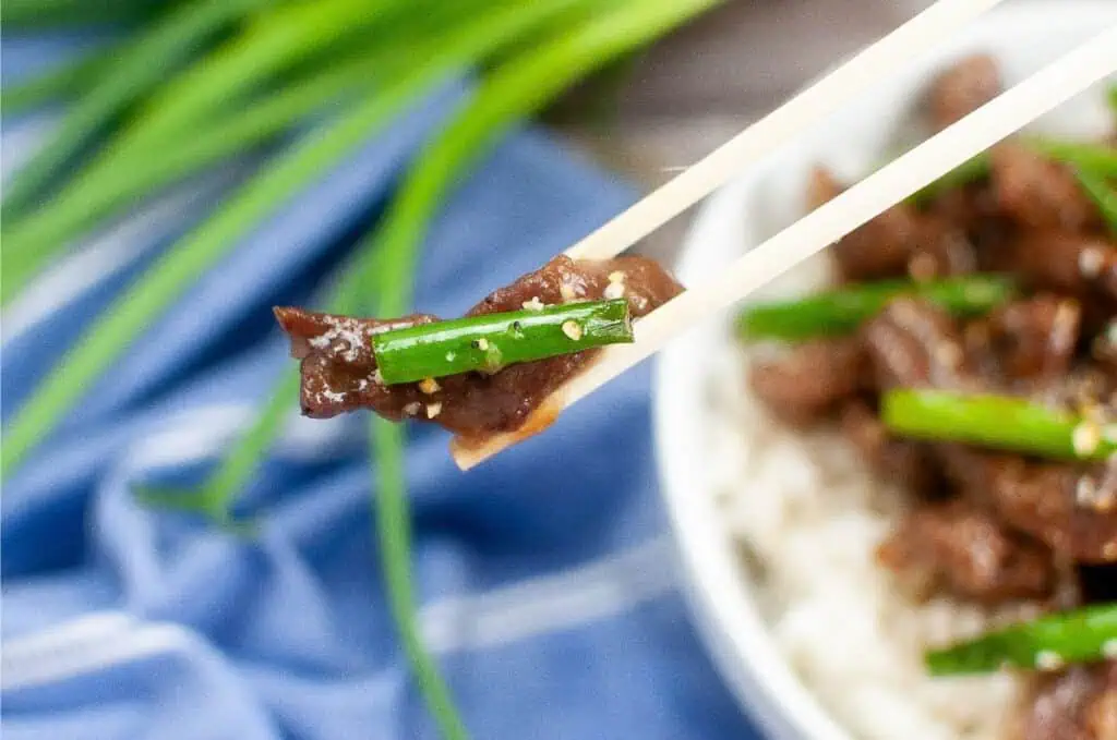 Chopsticks holding a piece of Mongolian Beef and green onion hover above a bowl of rice, with fresh green onions in the background.